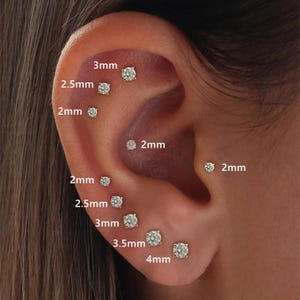 Puede incluir: Primer plano de una oreja con múltiples pendientes de botón dorados. Los pendientes varían en tamaño de 2 mm a 4 mm, cada uno con una gema redonda y transparente. La imagen destaca las joyas.
