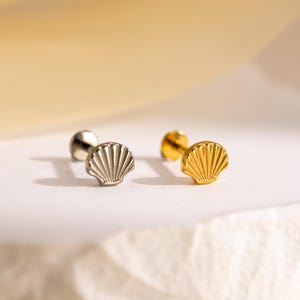 May include: Close-up of four small seashell-shaped earrings. Two are silver-colored, and two are gold-colored. The earrings are on a white surface with a soft yellow background.