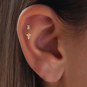 May include: A gold-colored earring with a dangling cross design, adorned with small, clear gemstones. The earring is attached to a rectangular stud. The image shows the earring on an ear.