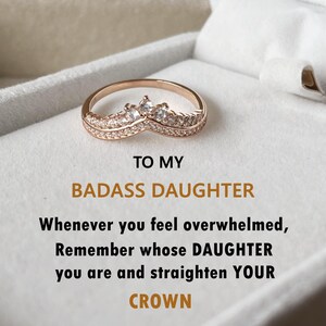 May include: A gold ring with a crown design and small clear stones. The ring is in a white box with the text "TO MY BADASS DAUGHTER Whenever you feel overwhelmed, Remember whose DAUGHTER you are and straighten YOUR CROWN".