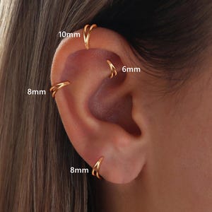 Puede incluir: Pendientes de aro dorados en varios tamaños, incluyendo 6 mm, 8 mm y 10 mm. Los pendientes se muestran en una oreja, destacando sus diferentes ubicaciones y estilos. La imagen muestra el diseño de los pendientes.