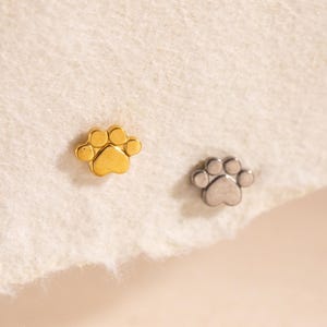 以下が含まれることがあります： 小さな足跡型のスタッドピアス2つ。1つは金色、もう1つは銀色です。ピアスは明るい色のテクスチャのある表面にあります。