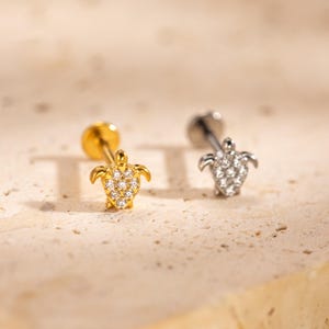 May include: Two stud earrings featuring a turtle design. One earring is gold-toned with clear stones, the other is silver-toned with clear stones. The earrings are on a textured, light-colored surface.