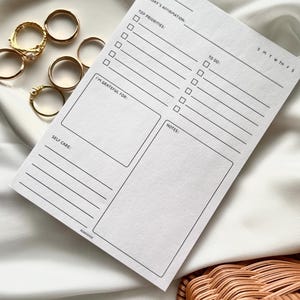 May include: A white printable daily planner with sections for date, today's affirmation, top priorities, to do, I'm grateful for, notes, and self care. The planner also includes a small calendar with the days of the week.