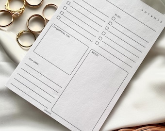 May include: A white printable daily planner with sections for date, today's affirmation, top priorities, to do, I'm grateful for, notes, and self care. The planner also includes a small calendar with the days of the week.