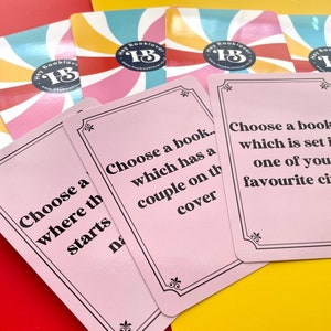 May include: Pink cards with black borders and text. The text on the cards reads "Choose a book... which has a couple on the cover" and "Choose a book which is set in one of your favourite cities".