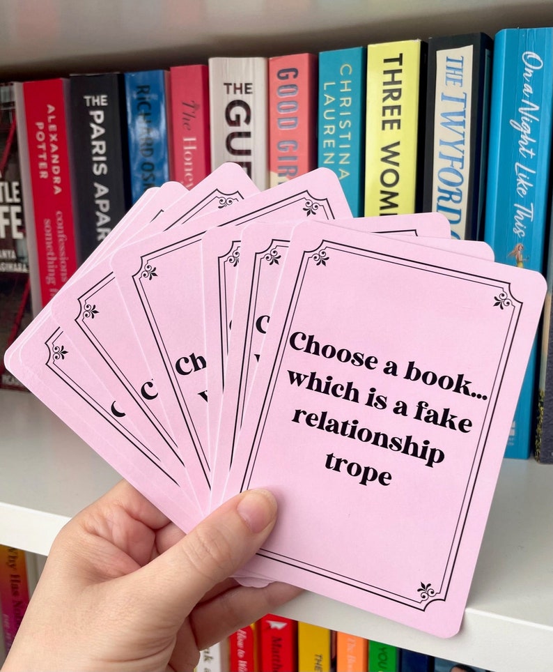 May include: Pink cards with black borders and text. The text on the top card reads "Choose a book... which is a fake relationship trope".