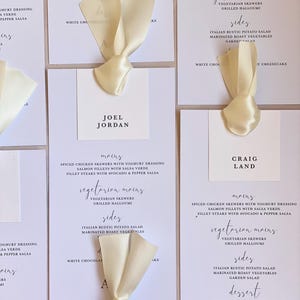 May include: Wedding menu cards with ivory satin ribbon. Each card displays menu details and guest names, like "Joel Jordan" and "Craig Land", in a minimalist style. The cards list mains, sides, and dessert options.