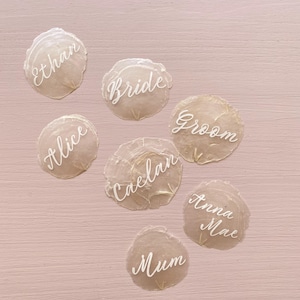 May include: A set of seven white calligraphy place cards on a light pink background. The cards are written with the words "Bride", "Groom", "Ethan", "Alice", "Caelan", "Mum", and "Anna Mae".