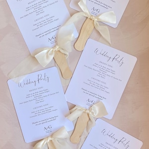 May include: White wedding fan with the text "Wedding Party" in elegant script. The fan lists the bridal party and groom's side details. Each fan has a wooden handle and a cream-colored ribbon.