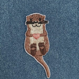 Könnte beinhalten: Gestickter Aufnäher mit einem Cartoon-Otter, der ein rosa Herz hält. Der Otter ist braun und beige mit schwarzen Schnurrhaaren und Augenbrauen. Der Aufnäher befindet sich auf einem blauen Jeans-Hintergrund, mit dem Etsy-Logo oben.
