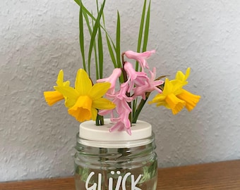 Op de afbeelding: Een kleine glazen pot met het woord "GLÜCK" in witte letters, met een bloemstuk. Het arrangement bevat gele narcissen, roze hyacinten en groen gras. De pot staat op een houten oppervlak.