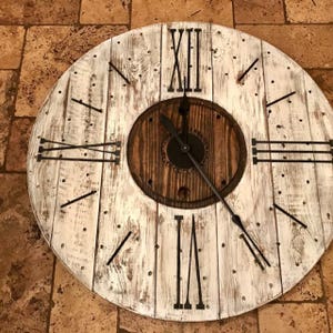 Puede incluir: Un reloj grande de madera blanqueada con números romanos y manecillas de metal negro. La esfera del reloj está hecha de un carrete de madera reutilizado.