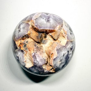 May include: A spherical amethyst geode with a rough, textured surface. The geode features a mix of purple and brown hues, with crystalline structures visible throughout. The overall shape is round, and the geode is set against a white background.