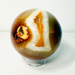 May include: A polished agate sphere, showcasing a blend of earthy tones. The orb features bands of brown, orange, and white, with a crystalline center. It rests on a clear, circular stand, highlighting its natural beauty.