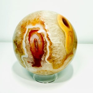 May include: A polished agate sphere with a mix of colors, including shades of orange, brown, and white. The stone has a unique pattern with a central red and white design. It sits on a clear, round stand.