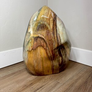 May include: A large, polished, freeform stone sculpture featuring a smooth, glossy surface with swirling patterns of earthy colors including deep brown, golden yellow, cream, and muted green, creating a natural, organic, and multi-toned appearance.