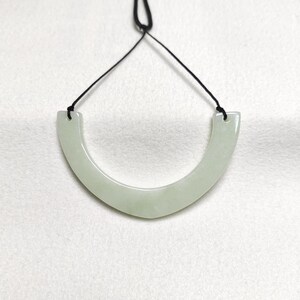Puede incluir: Un collar de jade verde pálido con un colgante curvo en forma de U. El colgante está suspendido de un cordón negro. El jade tiene una superficie lisa y pulida y una calidad sutil y translúcida.
