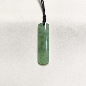 Puede incluir: Un colgante de jade verde con forma cilíndrica, suspendido de un cordón negro. El jade tiene un aspecto moteado con diferentes tonos de verde. El colgante se muestra sobre un fondo blanco.