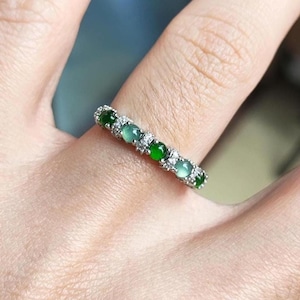 May include: A silver ring adorned with green gemstones and small clear stones. The ring showcases a delicate design with alternating dark and light green stones. The ring is worn on a finger. The gemstones are likely measured in millimetres.