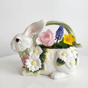 May include: A white ceramic bunny planter with a green handle, decorated with white daisies and a pink rose. The planter holds a pink tulip, blue grape hyacinth, and yellow flower, perfect for spring decor.