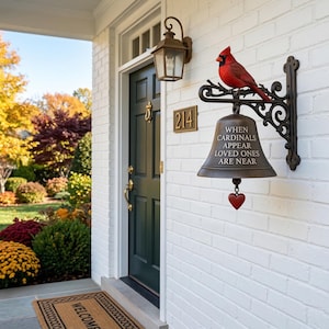 Puede incluir: Una campana decorativa montada en la pared con un cardenal rojo en la parte superior. La campana es de color bronce y tiene el texto "WHEN CARDINALS APPEAR LOVED ONES ARE NEAR". Un pequeño corazón rojo cuelga debajo.