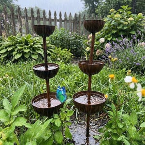 May include: Two tiered, rustic brown metal fountains with water cascading down into the bowls. A blue and green butterfly rests on one of the bowls. The fountains are in a garden setting with green plants and flowers.