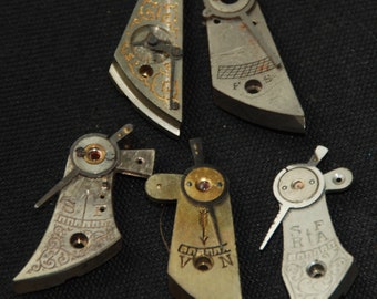 May include: Five antique watch parts with intricate designs and markings. The parts are made of metal and have a silver and gold finish. Some of the parts have numbers and letters engraved on them.