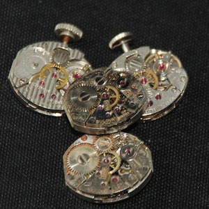 May include: Close-up of four vintage watch movements, showcasing intricate gears, jewels, and metal components. The Bulova brand is visible on one movement. The watch parts are silver and gold in color, with red jewels.