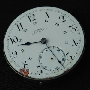 May include: A vintage Regina pocket watch face with a white dial, black numerals, and blue hands. The watch face includes a smaller subdial and the text "REGINA J.U. MUNNS MOOSE JAW, SASK". The watch face has some wear and tear.