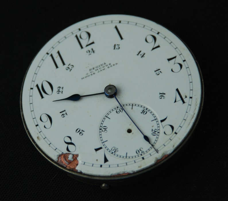 May include: A vintage Regina pocket watch face with a white dial, black numerals, and blue hands. The watch face includes a smaller subdial and the text "REGINA J.U. MUNNS MOOSE JAW, SASK". The watch face has some wear and tear.