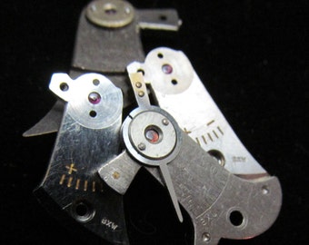 May include: Close-up of several small, silver-colored watch movement parts. Each piece has a circular design with a small, red jewel in the center. Some parts have markings and numbers etched on them. The background is black.