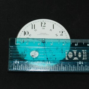 May include: A vintage watch face with black numerals on a white background, measuring approximately 2 inches in diameter. The face includes the text "REGINA J. MUNKEE MOOSE JAW SASK". A clear ruler is placed across the face.