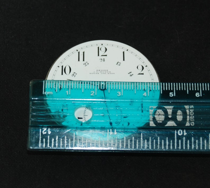May include: A vintage watch face with black numerals on a white background, measuring approximately 2 inches in diameter. The face includes the text "REGINA J. MUNKEE MOOSE JAW SASK". A clear ruler is placed across the face.