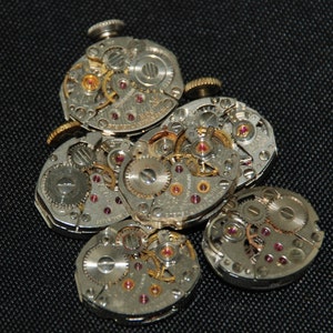 May include: Close-up of several silver watch movements with intricate gears and ruby jewels. The watch parts are arranged in a cluster on a black surface.