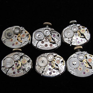 Puede incluir: Seis movimientos de reloj plateados con engranajes intrincados y rubíes. Los movimientos están etiquetados con la marca "Bulova" y el número de rubíes.