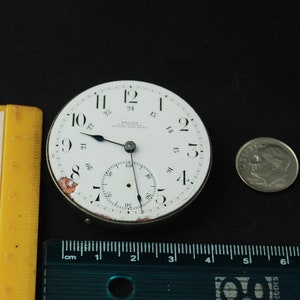 May include: A vintage watch face with a white dial, black numerals, and blue hands. The watch face has a subdial and the text "REGINA WATCH CO. NEW YORK". A dime is next to the watch face for size comparison. A yellow ruler is on the left.