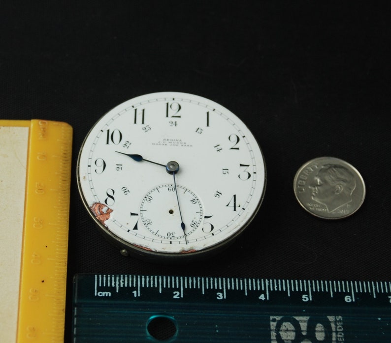May include: A vintage watch face with a white dial, black numerals, and blue hands. The watch face has a subdial and the text "REGINA WATCH CO. NEW YORK". A dime is next to the watch face for size comparison. A yellow ruler is on the left.