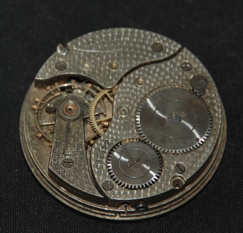 May include: Close-up of a vintage watch movement. The silver-toned mechanism features intricate gears, screws, and a balance wheel. The word "REGIN" is engraved at the top. The serial number "3483655" is visible.