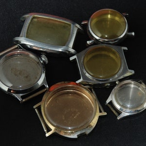 May include: A selection of vintage watch cases in different shapes and sizes. The cases are made of silver-toned metal and have empty interiors. Some cases are round, while others are rectangular. The watch cases are arranged on a black surface.