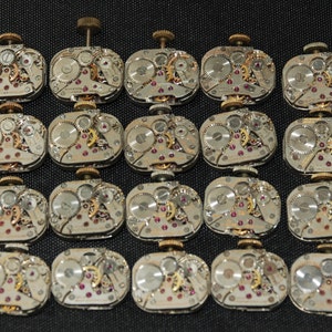 Puede incluir: Veinte movimientos de reloj antiguos con engranajes intrincados y rubíes. Los movimientos son plateados y tienen una forma circular.