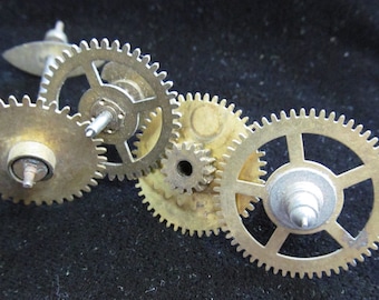 May include: Close-up of four interlocking brass gears with a silver axle in the center. The gears are arranged in a circular pattern and have a worn, antique look.