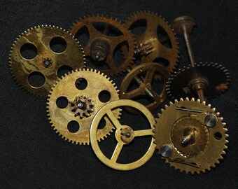 May include: A collection of vintage brass clock gears of various sizes and designs. The gears have a worn, aged appearance, with visible wear and tear. Some gears have small holes, while others have intricate teeth. The gears are arranged on a dark background.