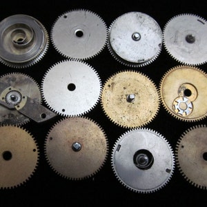 May include: A selection of vintage watch components, including gears, springs, and other mechanical parts. The gears are of various sizes and materials, with silver and brass hues. The parts are arranged on a black backdrop, highlighting the intricate details.