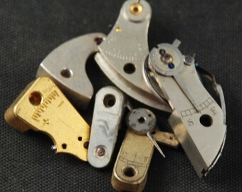 May include: A collection of small, intricate watch parts in various shapes and metallic finishes. Components include silver, gold, and brass pieces, some with markings and tiny screws. The parts are scattered against a dark background.