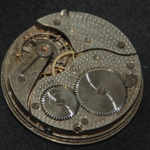 May include: Close-up of a vintage watch movement. The silver-toned mechanism features intricate gears, a balance wheel, and the word "REGINA" engraved on the plate. The design includes a series of small screws and a textured surface.