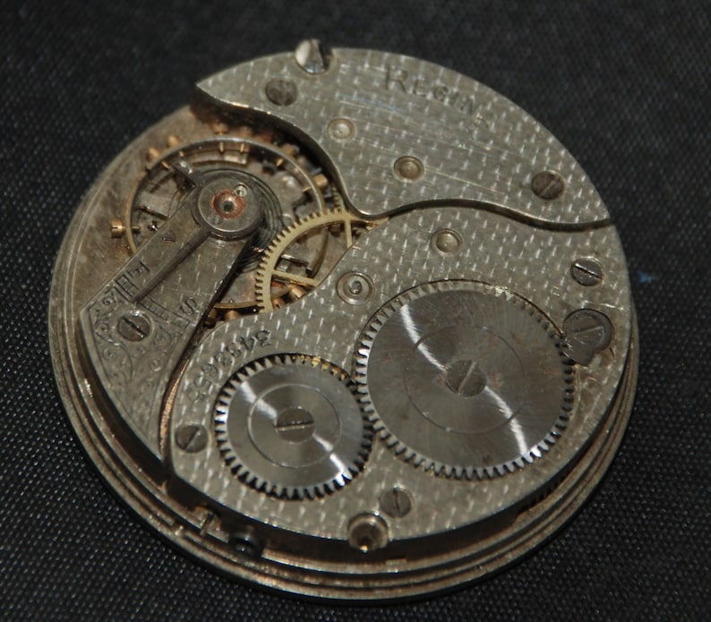 May include: Close-up of a vintage watch movement. The silver-toned mechanism features intricate gears, a balance wheel, and the word "REGINA" engraved on the plate. The design includes a series of small screws and a textured surface.