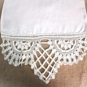 May include: Close-up of a white fabric with a detailed, crocheted lace border. The lace features a repeating floral pattern along the top edge and a diamond-shaped openwork design at the bottom, creating an elegant, handcrafted look.