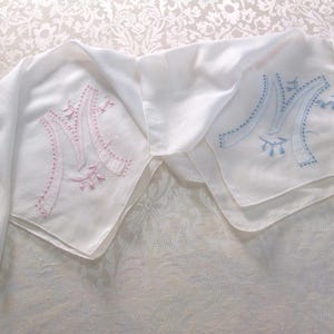May include: Two white handkerchiefs with embroidered capital letter "M" designs. One handkerchief features a pink "M" with floral accents, while the other has a blue "M" with similar detailing. The handkerchiefs are displayed on a patterned white surface.