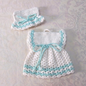 May include: White crocheted baby dress with a blue trim and matching hat. The dress has a bow at the waist.
